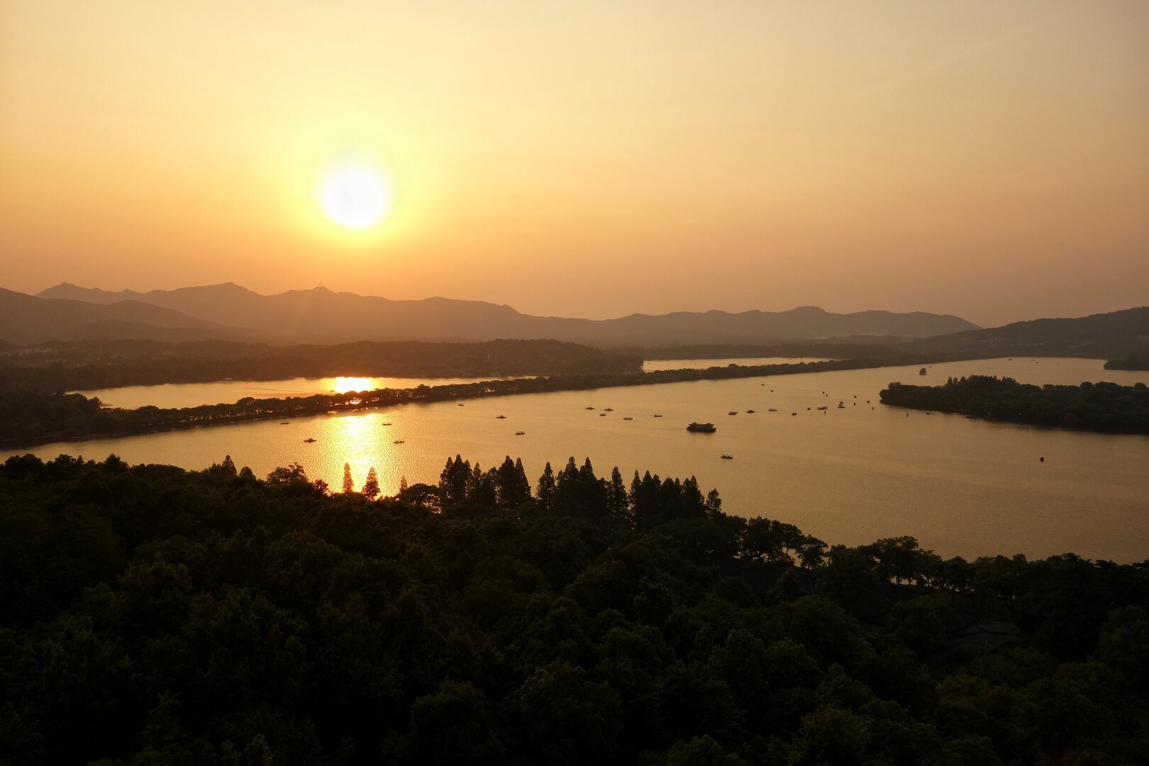 Golden sunset over West Lake in Hangzhou, China, a famous UNESCO World Heritage site. The large sun hangs low above distant hazy mountain ridges, casting warm amber and orange light across the calm, golden-hued waters. The expansive lake stretches towards the horizon, dotted with numerous small boats and tree-covered islands, their reflections shimmering in the golden glow. A dense belt of pine trees in the foreground creates dark silhouettes, contrasting sharply with the luminous sky. Soft mist hangs over the water, adding to the atmospheric beauty. Layer upon layer of mountains are silhouetted on the far horizon, creating a sense of depth. Gentle ripples spread from the boats across the otherwise mirror-like surface. The serene scene captures the peaceful transition from day to evening, with the entire landscape bathed in warm, golden light.