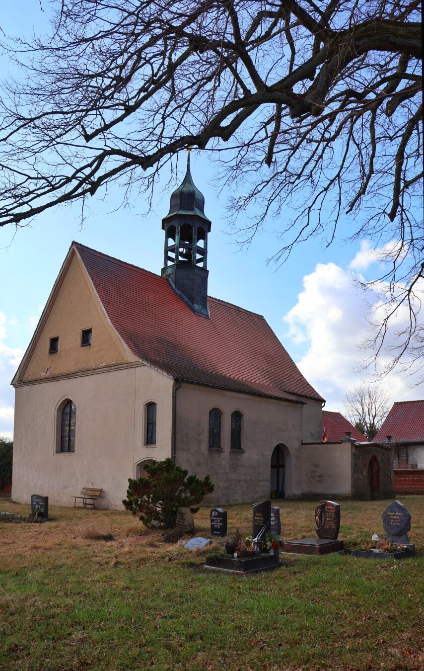 A small village church in Klein Glien with light-coloured rendered walls and a red terracotta tiled gable roof, featuring an integrated bell tower with a pointed top. The church has arched windows, small square windows near the roof, and a main arched entrance. In the foreground is a churchyard cemetery with numerous gravestones and tombstones of varying sizes and designs. A bare deciduous tree with spreading branches frames the top of the scene, while a small conifer and a wooden bench sit near the church. The sky is blue with white clouds, and other red-roofed buildings are visible in the background.