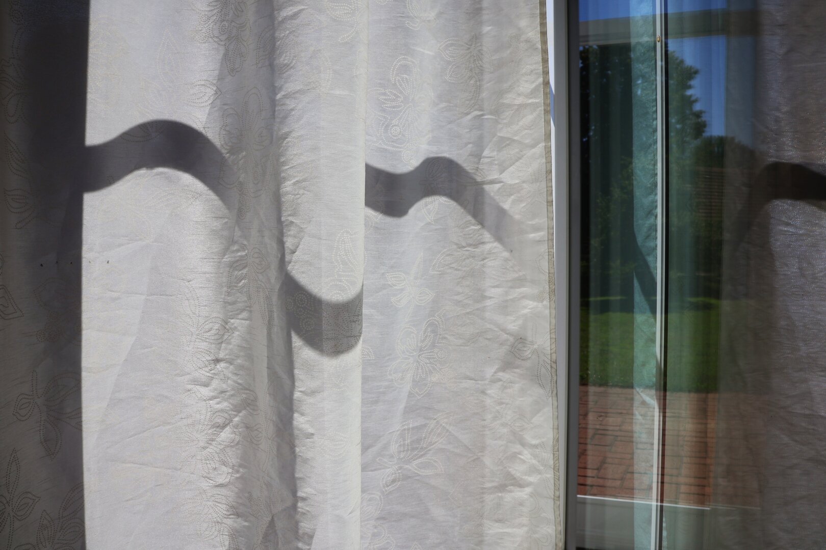 An open patio door with white, textured curtains, partially drawn. The folds of the curtains create dark, wavy shadows on the fabric, forming the silhouette of a smiling face with curved lines for eyes and an upturned mouth. Through the glass panes, the vibrant greenery of an outdoor garden with a red brick patio, trees, and grass is visible.