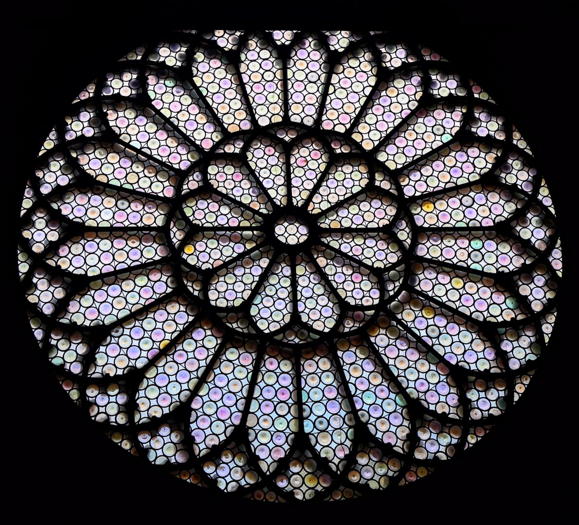 Circular stained glass rose window from Trieste Cathedral, featuring an intricate geometric design composed of numerous small circular panes of iridescent glass in soft pastel hues. Dark lead caming divides the window into radial and concentric patterns, creating a symmetrical, kaleidoscopic effect with subtle colour variations against the black background.