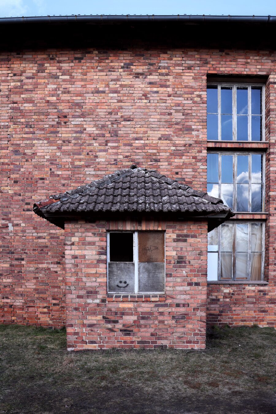 Red-brick building featuring a small, boarded-up window surmounted by a triangular, moss-covered tiled roof. A smiley face is drawn on the lower pane. To the right, two tall, wooden-framed windows with weathered glass and peeling paint reflect a blue sky with clouds. The structure has a roof and sits on a patch of grass and bare ground.