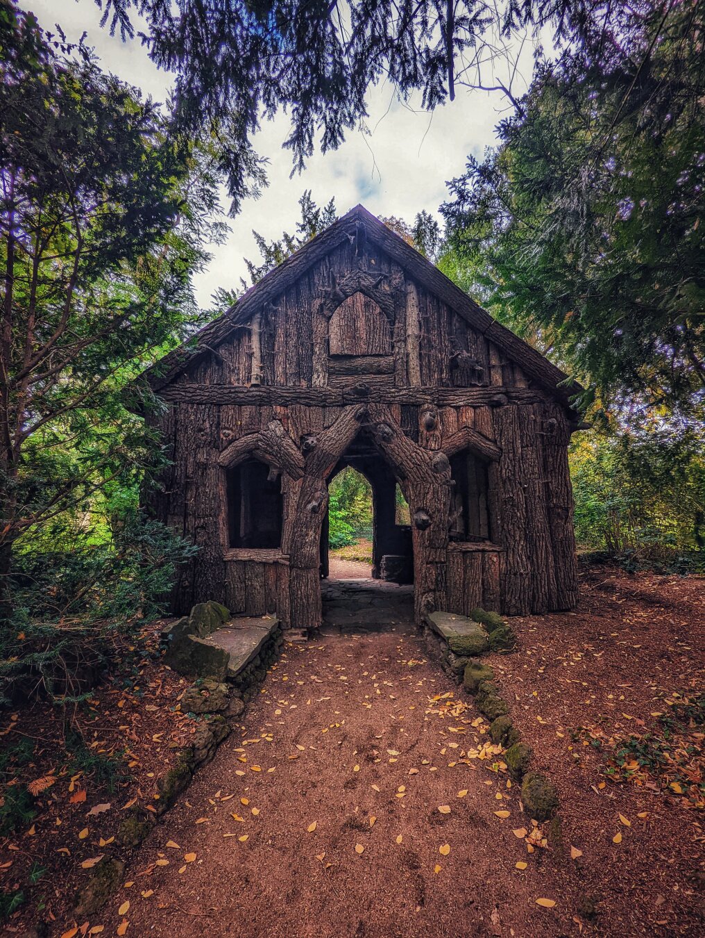 A charmingly rustic Borkenhäuschen (bark house) in Oranienbaum-Wörlitz, Germany, captured on a slightly overcast day. The small, wooden structure is fashioned from gnarled logs and branches, giving it a fairy-tale or witch-house aesthetic. A dirt path, scattered with autumn leaves, leads up to its central arched entrance. Two small, rustic benches made of stone and moss sit on either side of the entrance. The house is nestled amongst dense green trees and foliage, with dappled light filtering through the leaves above, enhancing its whimsical and secluded feel. A charmingly rustic Borkenhäuschen (bark house) in Oranienbaum-Wörlitz, Germany, captured on a slightly overcast day. The small, wooden structure is fashioned from gnarled logs and branches, giving it a fairy-tale or witch-house aesthetic. A dirt path, scattered with autumn leaves, leads up to its central arched entrance. Two small, rustic benches made of stone and moss sit on either side of the entrance. The house is nestled amongst dense green trees and foliage, with dappled light filtering through the leaves above, enhancing its whimsical and secluded feel.