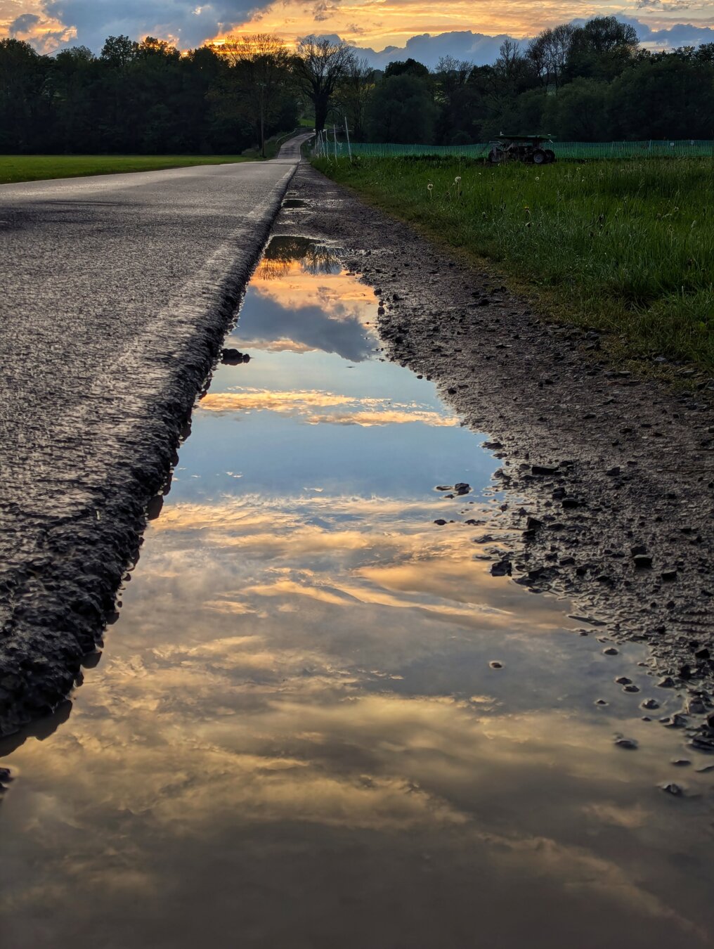 A serene, low-angle shot captures a long puddle along the side of a country road at dusk. The sky, painted with soft orange and grey clouds, is perfectly reflected in the still water of the puddle, creating a mirror image. The tarmac road stretches into the distance on the left, while a grassy verge and a field, with hints of agricultural machinery, line the right. A dark band of trees forms the horizon, silhouetted against the colourful twilight sky. A serene, low-angle shot captures a long puddle along the side of a country road at dusk. The sky, painted with soft orange and grey clouds, is perfectly reflected in the still water of the puddle, creating a mirror image. The tarmac road stretches into the distance on the left, while a grassy verge and a field, with hints of agricultural machinery, line the right. A dark band of trees forms the horizon, silhouetted against the colourful twilight sky.