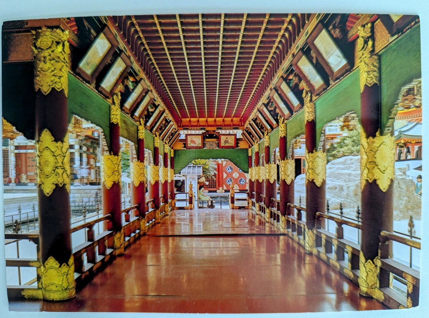 A postcard of a low-angle, eye-level shot shows a covered walkway with polished wooden flooring, flanked by ornate dark brown pillars embellished with gold leaf carvings. The pillars support a dark brown, coffered ceiling with panels of red and gold detailing. Natural light streams in from the open sides of the walkway, illuminating the polished floor.
On either side, wooden railings run along the length of the walkway. Beyond the railings, glimpses of buildings with light-coloured walls and dark roofs can be seen. At the far end of the walkway, an arched opening reveals a brightly coloured structure with red, white, and blue elements.
The overall impression is one of traditional East Asian architecture, showcasing intricate woodwork and decorative elements.