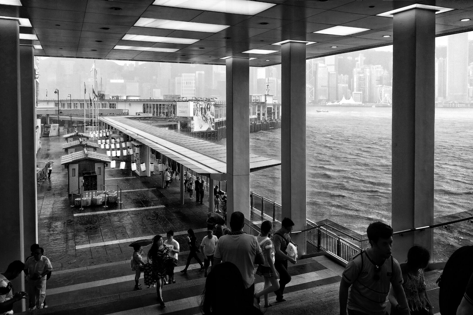 A black-and-white photograph captures Harbour City Ocean Terminal in Hong Kong during heavy rain. In the foreground, people walk under a covered walkway with tall columns, while steps descend towards the pier. The terminal building extends into the harbour, with calm water visible to the right. In the distance, Hong Kong’s dense skyline of towering skyscrapers looms through the downpour, creating a dramatic, atmospheric contrast.