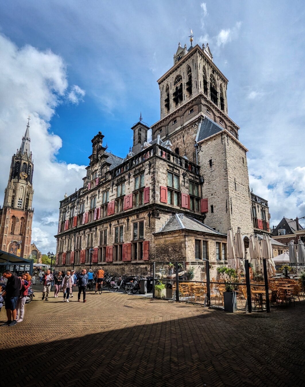 The historic Town Hall of Delft, Netherlands, stands prominently under bright daylight, its tall clock tower featuring ornate Gothic architectural details and crenellated parapets. The building's light grey stone facade showcases multiple rows of windows adorned with bright red shutters and steep gabled roofs. To the left, the tall, pointed church spire of Nieuwe Kerk with intricate stonework pierces the partly cloudy blue sky with scattered white clouds. In the foreground, a cobblestone square hosts outdoor cafe seating with wooden tables, chairs, and closed umbrellas, while people walk, chat, and gather in the lively plaza.