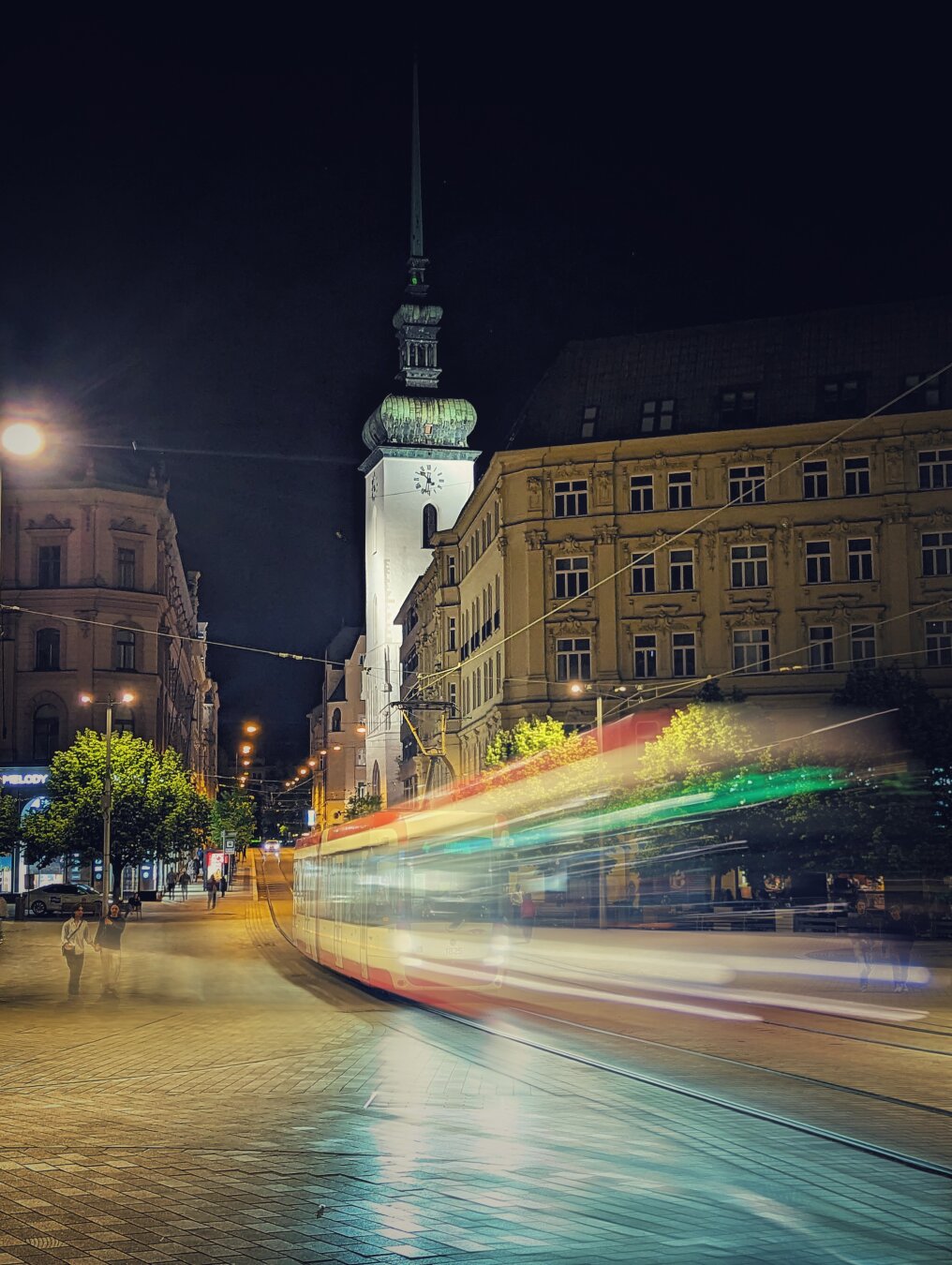 Long-exposure night photograph of a city street featuring a tall, illuminated clock tower with a spire and clock face, flanked by ornate buildings. A moving tram creates vibrant red, green, and white light trails across the scene. Cobblestone pavement, street lights, and blurred pedestrians contribute to the lively urban atmosphere.