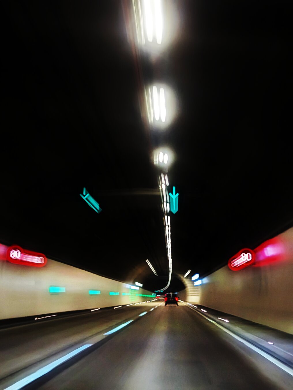 A motion-blurred night-time view from inside a car driving through a tunnel, with bright overhead lights creating streaks, green exit signs, and red 80 km/h speed limit signs on the walls.
