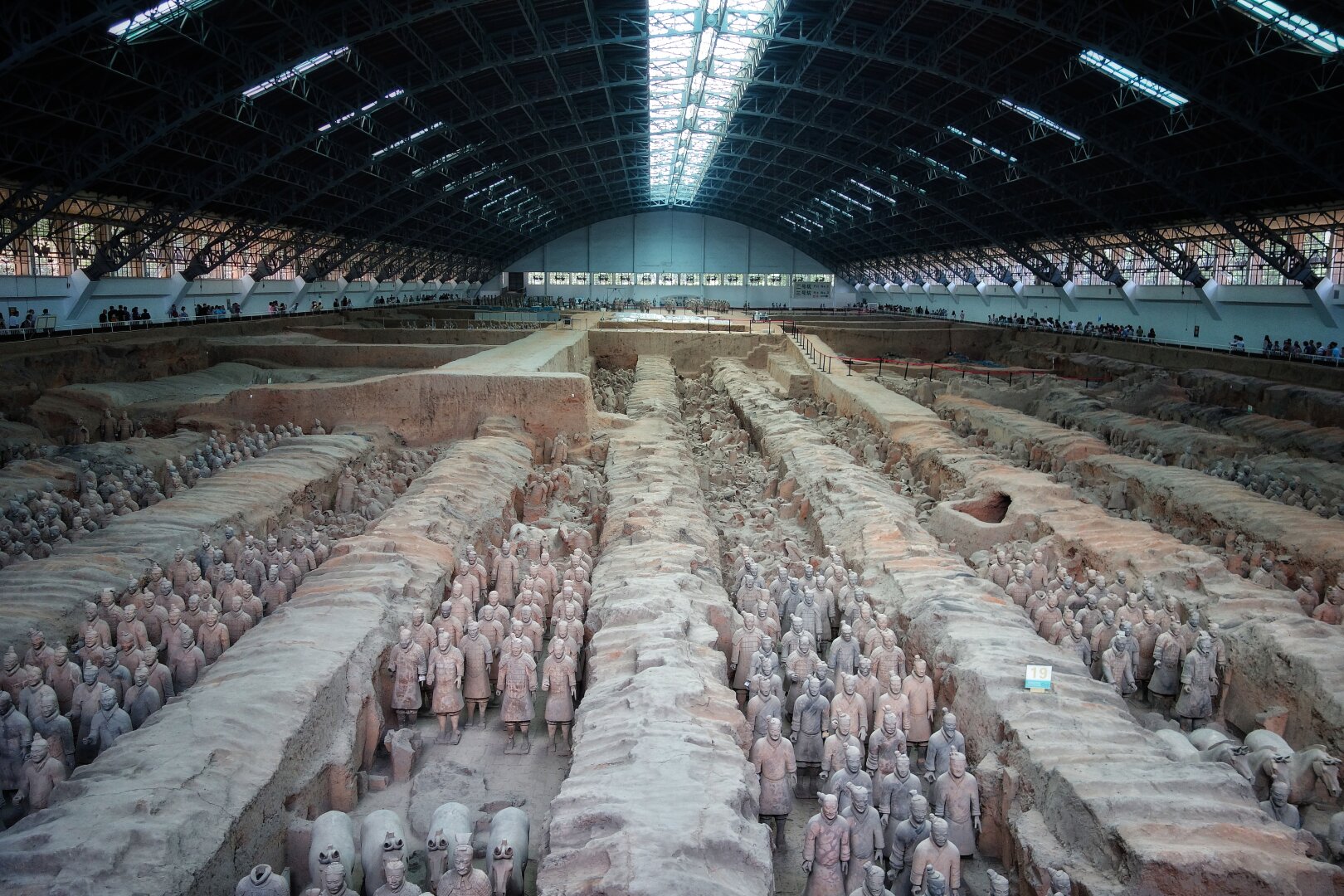 An aerial view of the Terracotta Army excavation site in Xi’an, featuring rows of life-sized terracotta warrior statues arranged in military formation, housed within a vast protective hangar with a metal framework roof.