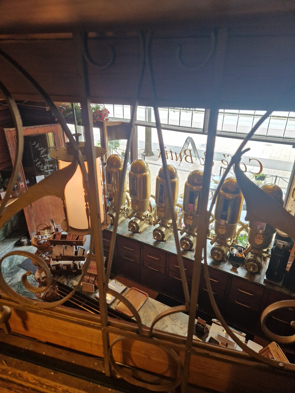 A view from an indoor balcony above the entrance of the café. In the background the very old coffee grinders in the shop window are visible. In the foreground, the scrollwork fence of the balcony.