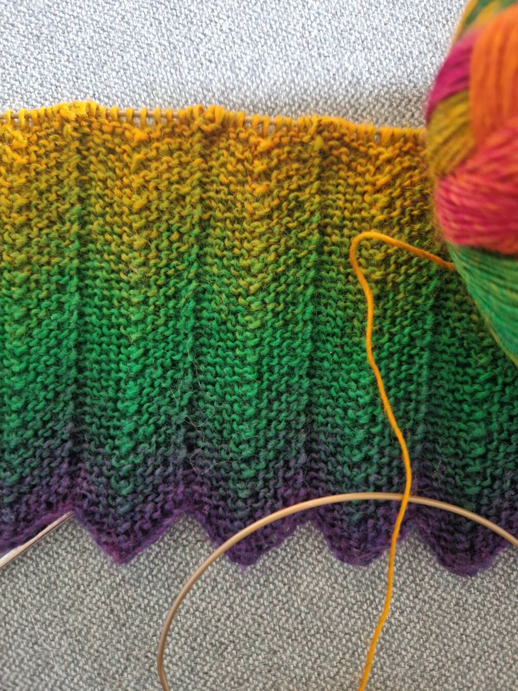 A close-up photo of a scarf in progress on circular knitting needles with the cable partly visible. The knitted fabric has a chevron-like pattern and displays a vibrant gradient of colors, transitioning smoothly from yellow at the top, through shades of green, ending with purple at the bottom. The ball of yarn is partly visible to the right, teasing soon to be added pink and orange colours. The whole piece rests on light gray textured sofa fabric.