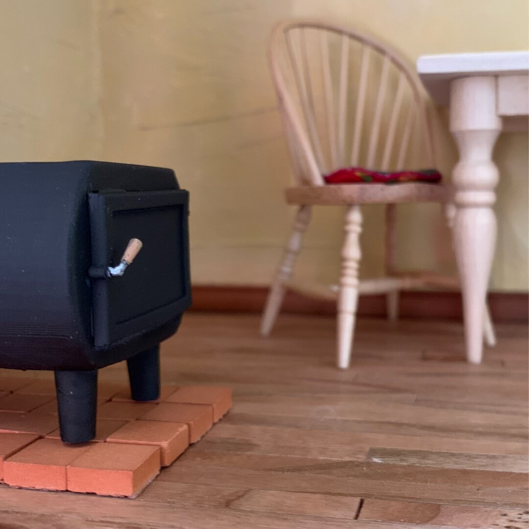 Miniature interior shown from a low angle near to the floor. It has cherry wood floors and light yellow plaster walls. At the left is a black wood stove sitting on red terracotta tiles. At center, against the back wall, is a Windsor chair with an embroidered chair pad. It is pushed slightly in at an angle under a white table, partially visible at the right side of the image.