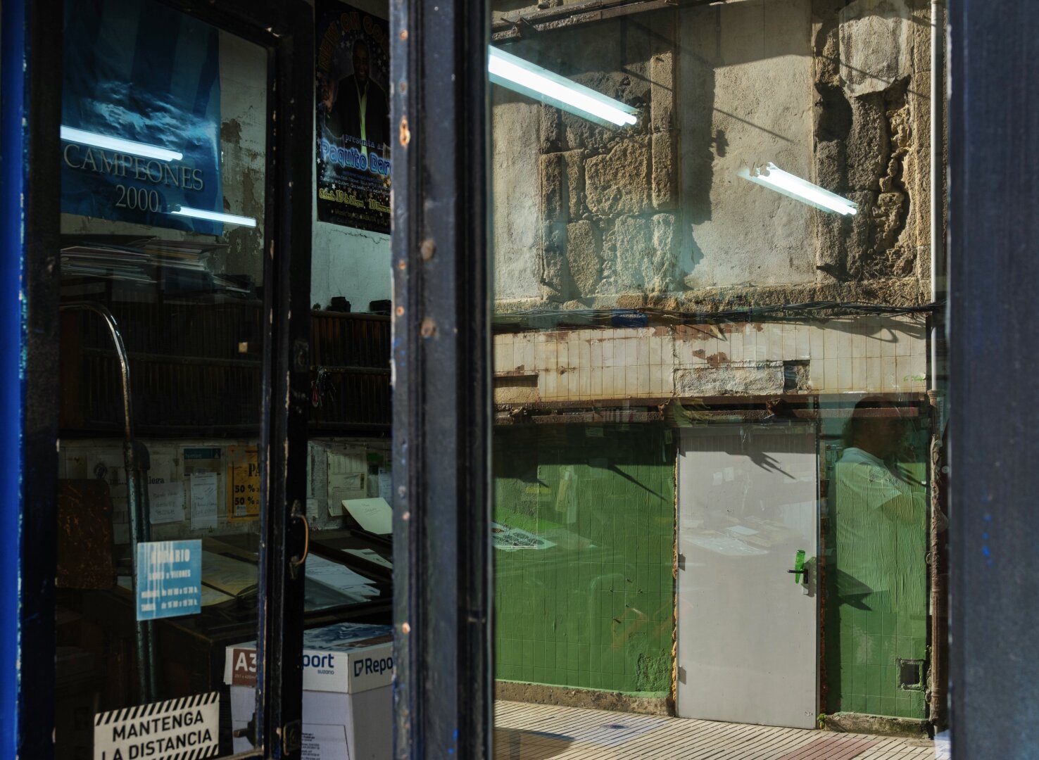 Das Bild zeigt eine Reflexion in einer Glastür, die einen Innenraum mit Stapeln von Papieren und einer metallischen Struktur zeigt, neben einer teilweise sichtbaren grünen Wand und einer weißen Tür im Hintergrund. Es gibt Leuchten oben und Plakate, die an der Außenwand zu sehen sind.