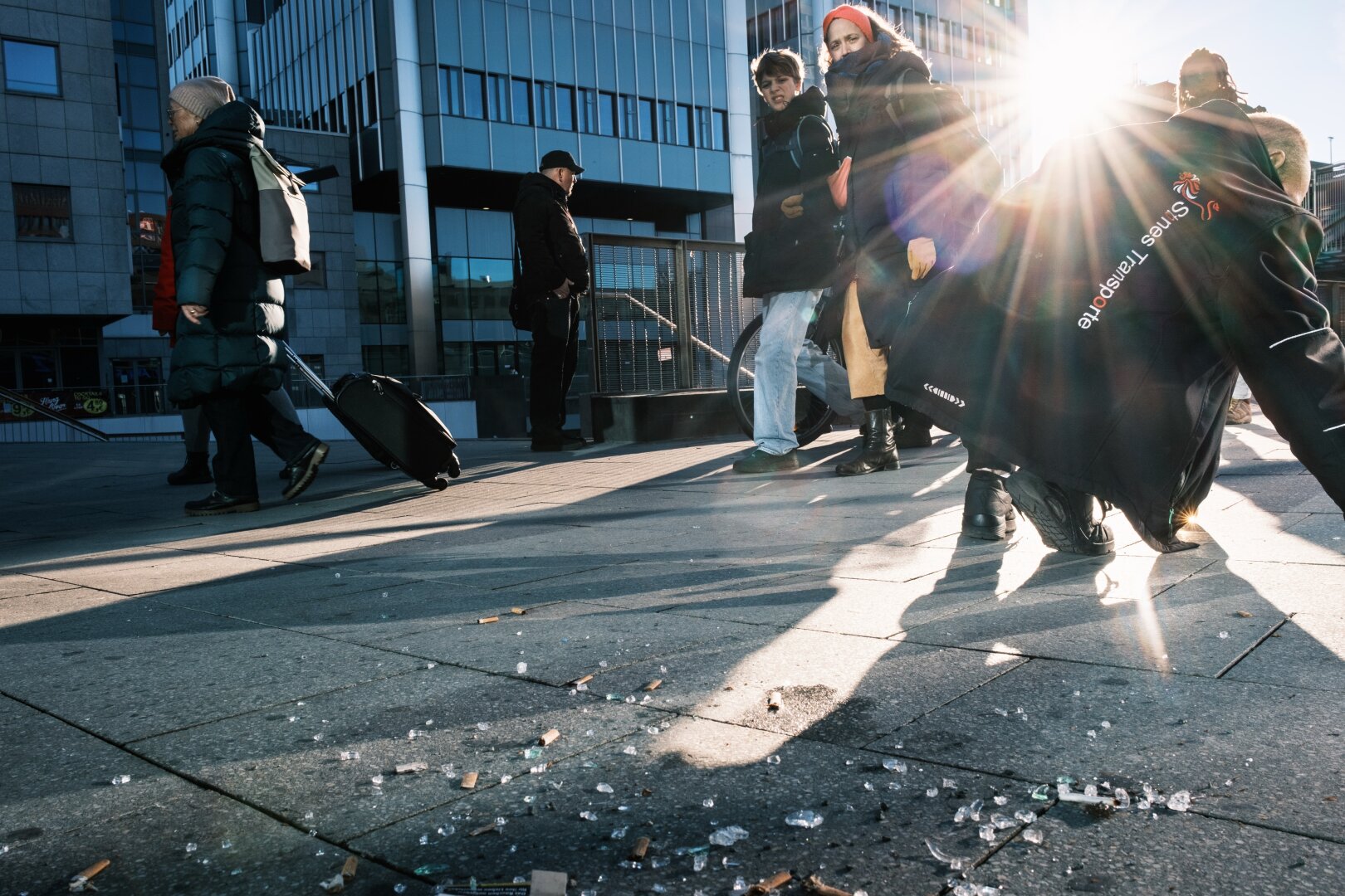 Eine geschäftige Stadtszene mit mehreren Menschen, die auf einem sonnenbeschienenen Bürgersteig gehen. Einige Personen tragen Gepäck oder Taschen, und auf dem Boden liegen sichtbare Abfälle, darunter Glasscherben und Zigarettenkippen. Im Hintergrund reflektieren moderne Gebäude das Sonnenlicht.