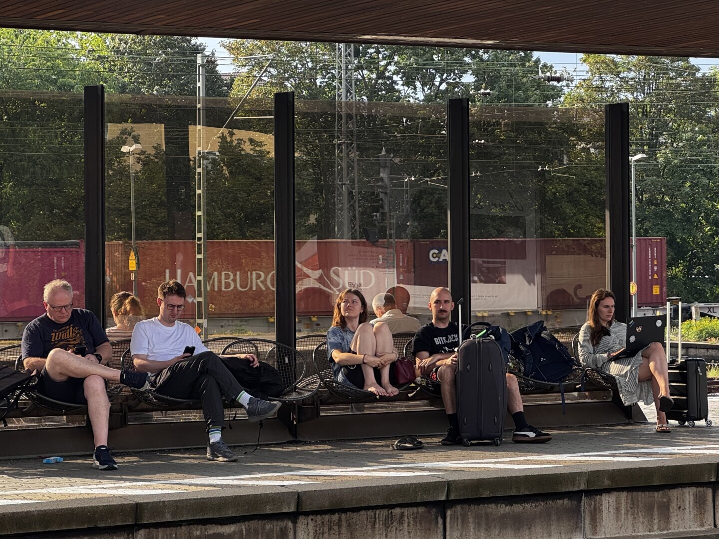 Eine Gruppe von Menschen sitzt auf einem Bahnsteig an einem Bahnhof; einige nutzen Smartphones und Laptops. In der Nähe steht Gepäck, im Hintergrund sind Bäume und ein Güterzug zu sehen. Die Szene ist sonnig und vermittelt eine entspannte Warteatmosphäre.