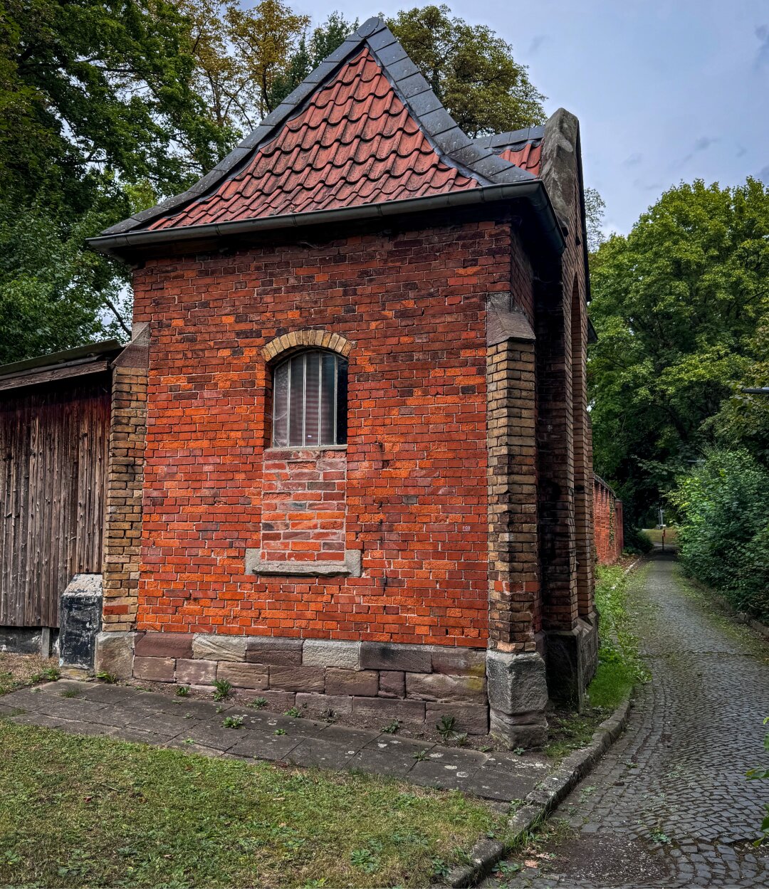 Ein kleines Backsteingebäude mit einem schrägen, rotgedeckten Dach, das neben einem von Grün umgebenen Kopfsteinpflasterwegs steht. Das Gebäude verfügt über ein einzelnes Bogenfenster und wirkt rustikal.