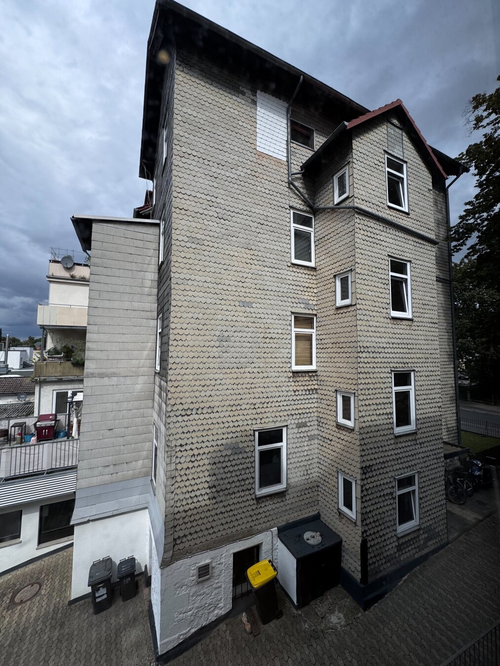 Ein mehrstöckiges Gebäude mit einer gemusterten Fassade, zahlreichen Fenstern und einem schrägen Dach. Im unteren Bereich stehen eine gelbe Mülltonne und ein dunkler Lagercontainer. Im Hintergrund ist ein bewölkter Himmel zu sehen.
