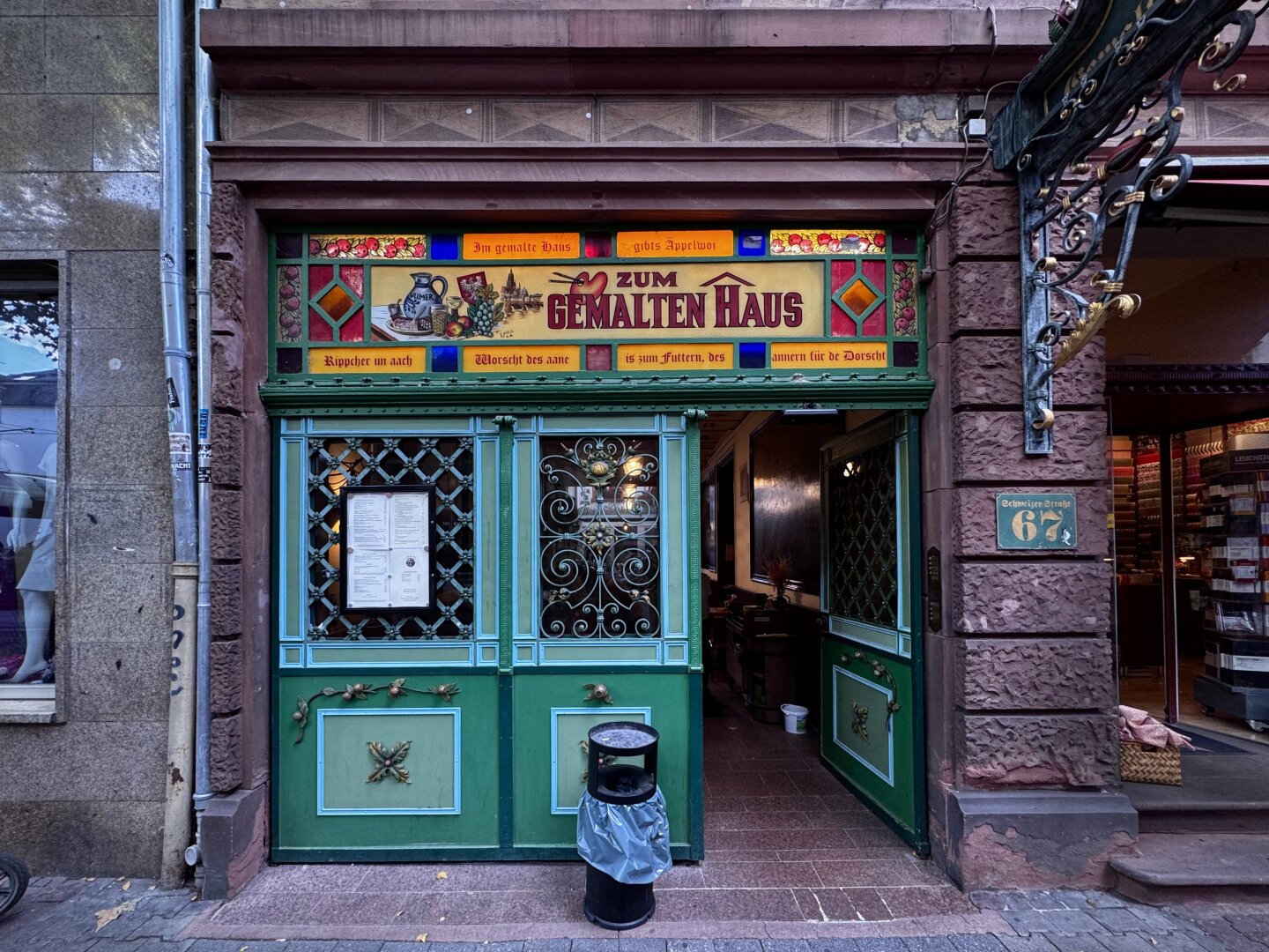 Der farbenfrohe Eingang zum Restaurant „Zum Gemalten Haus“ besticht durch dekorative Buntglasfenster und grüne Türen. Die Fassade weist kunstvolle Details auf, und neben der Tür ist die Speisekarte ausgehängt. Vor dem Eingang steht ein Mülleimer, und ein benachbartes Gebäude ist teilweise zu sehen.