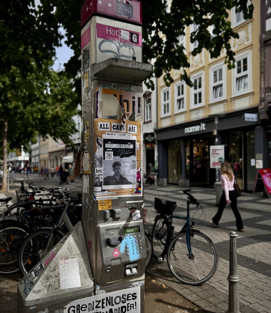 An einer Straße steht eine Telefonzelle, die mit verschiedenen Plakaten und Aufklebern beklebt ist. Auf einem Plakat steht „Grenzenloses Durcheinander“. In der Nähe stehen Fahrräder, und eine Person geht vorbei. Bäume spenden Schatten, während Gebäude und Geschäfte den Hintergrund bilden.