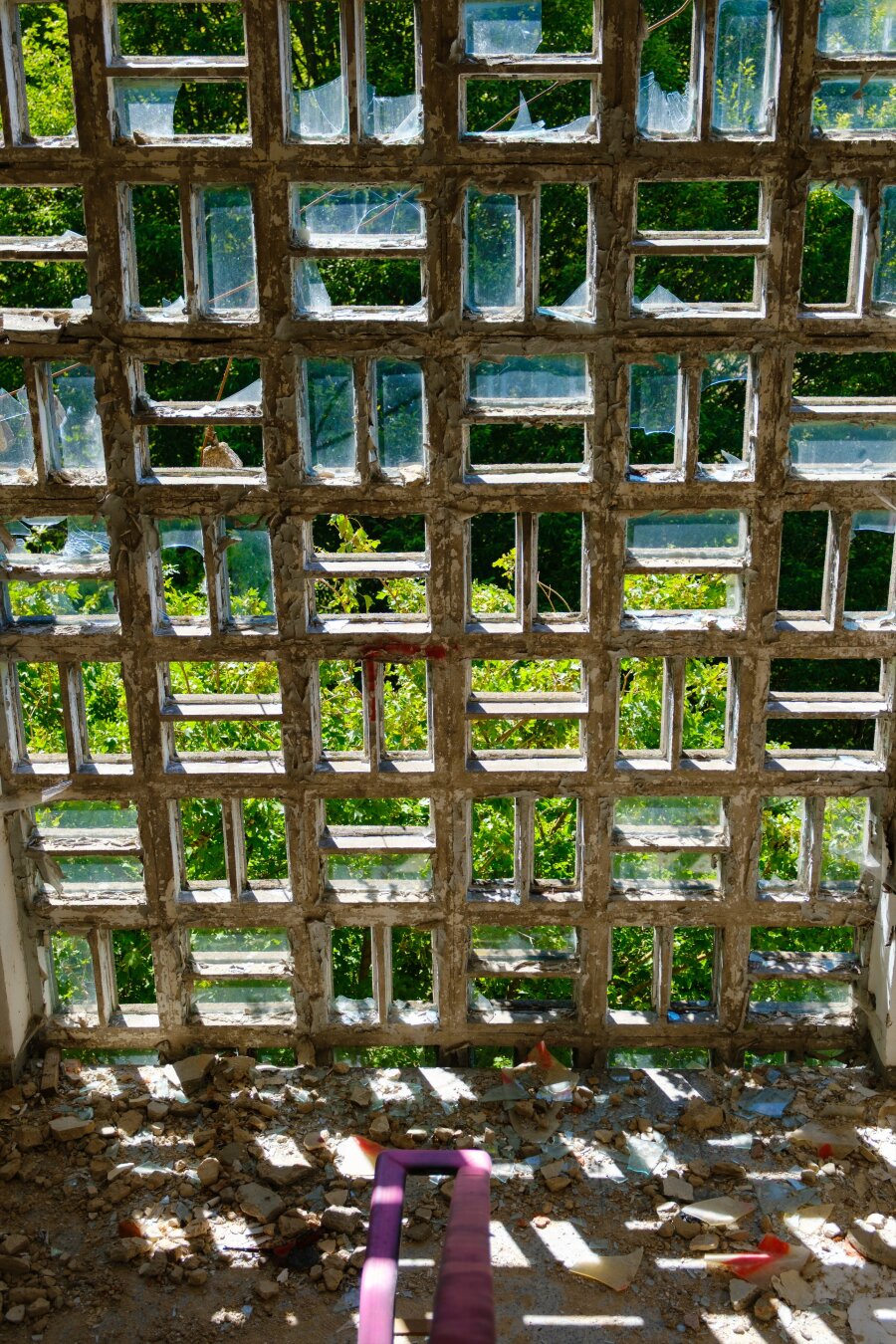 A deteriorating wall made of glass blocks with several broken panes, revealing greenery outside. Debris is scattered on the floor, and sunlight casts shadows through the openings.