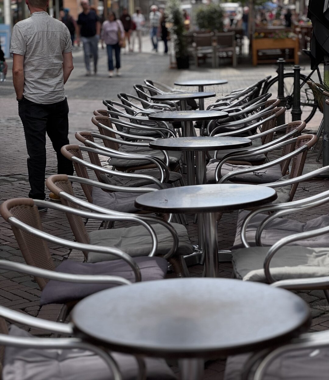 A man standing at the end of the row of metallic chairs in a café and staring anywhere.