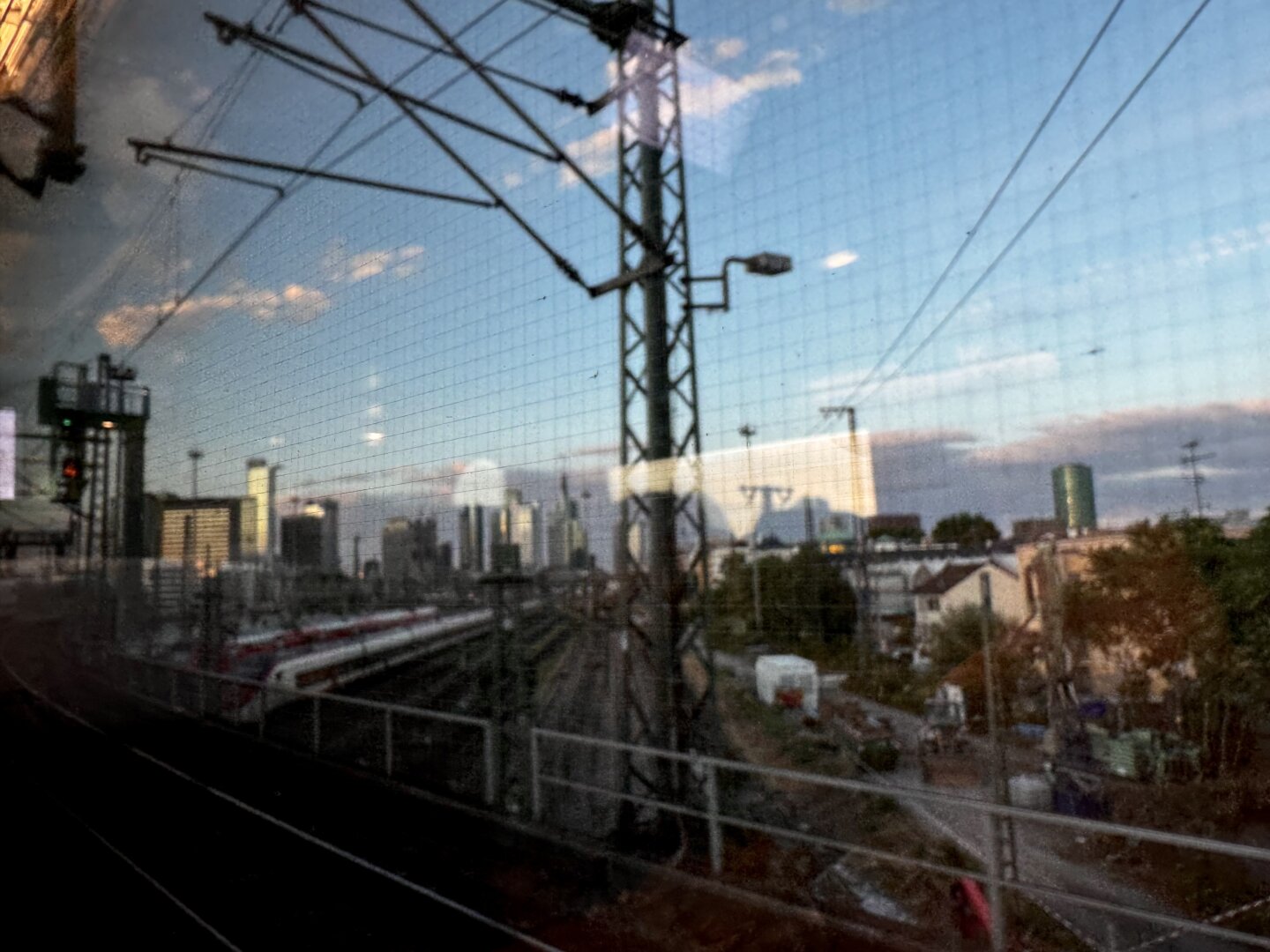 Ein unscharfer Blick aus einem Zugfenster auf die Skyline einer Stadt mit hohen Gebäuden, Stromleitungen und Bahngleisen. Im Vordergrund sind zudem ein Hauch von Grün und städtische Infrastruktur zu sehen.