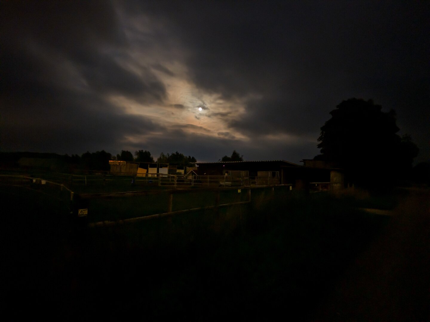 Eine schwach beleuchtete Bauernhofszene bei Nacht, in der ein Holzgebäude und ein Zaun unter einem bewölkten Himmel mit sichtbarem Mond zu sehen sind. Die Atmosphäre ist stimmungsvoll und rustikal, umgeben von dunkler Vegetation.