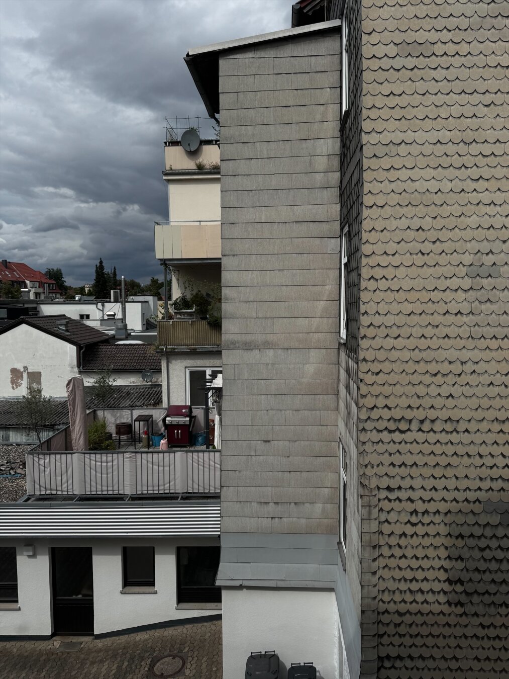 Ein Blick auf sich kreuzende Gebäudewände mit unterschiedlichen Strukturen und Materialien. Die linke Wand ist glatt und grau, während die rechte Wand ein wellenförmiges Muster aus Schindeln aufweist. Im Hintergrund sieht man einen bewölkten Himmel sowie einen flüchtigen Blick auf Dächer und einen Balkon