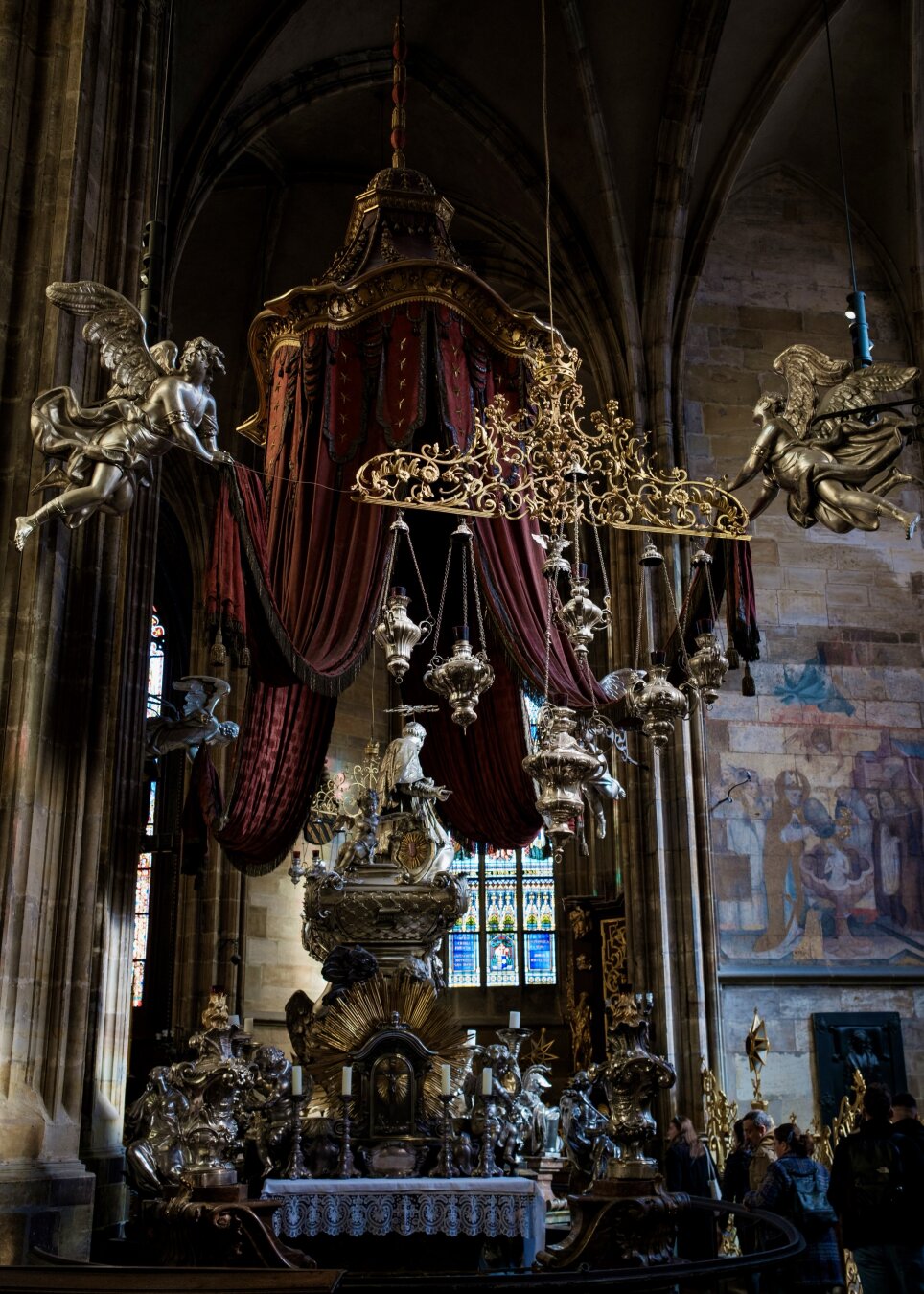 Das Innere des Prager Doms mit aufwendigen Silberverzierungen, darunter Engelsskulpturen, kunstvolle Kronleuchter und ein reiches rotes Baldachin. Buntglasfenster fügen buntes Licht hinzu, und die Besucher bewundern den überwältigenden Kitsch und katholischen Tinnef.