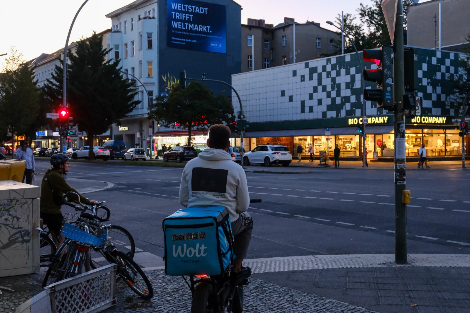 Zwei Fahrradfahrer warten an einer roten Ampel auf einer Kreuzung. Der vordere Radfahrer trägt einen großen, türkisfarbenen Lieferrucksack mit dem Logo von "Wolt", einem Essenslieferdienst. Beide Radfahrer sind von hinten zu sehen. Im Hintergrund sind Gebäude mit Geschäften, darunter ein "Bio Company"-Supermarkt, sowie eine beleuchtete Werbetafel mit dem Text "WELTSTADT TRIFFT WELTMARKT" zu erkennen. Die Straße ist belebt, mit Autos und Fußgängern. Die Atmosphäre wirkt lebendig und urban.