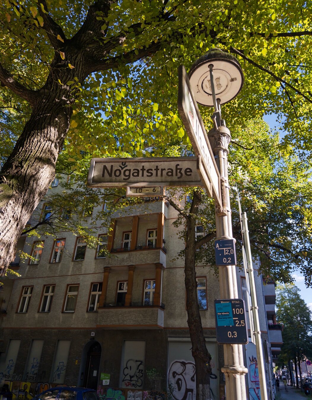 Ein Straßenschild für die Nougatstraße ist prominent ausgestellt, umgeben von hohen Bäumen mit grünen Blättern. Im Hintergrund ist ein Gebäude mit mehreren Fenstern und Graffiti zu sehen, zusammen mit nahe gelegenen Bushaltestellenschildern, die Informationen zum öffentlichen Verkehr anzeigen.