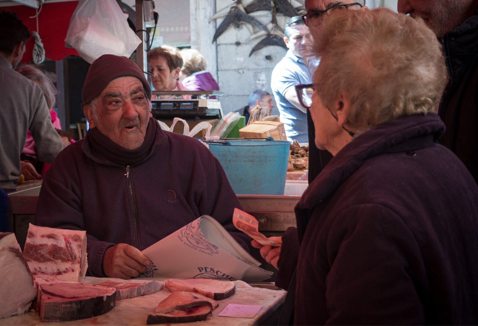 Ein älterer Mann und eine ältere Frau unterhalten sich an einem Marktstand, umgeben von Meeresfrüchten. Der Mann, der eine Mütze und eine dunkle Jacke trägt, wirkt lebhaft, während er ein Blatt Papier in der Hand hält. Auf dem Tisch vor ihm liegen verschiedene Fischstücke ausgestellt.