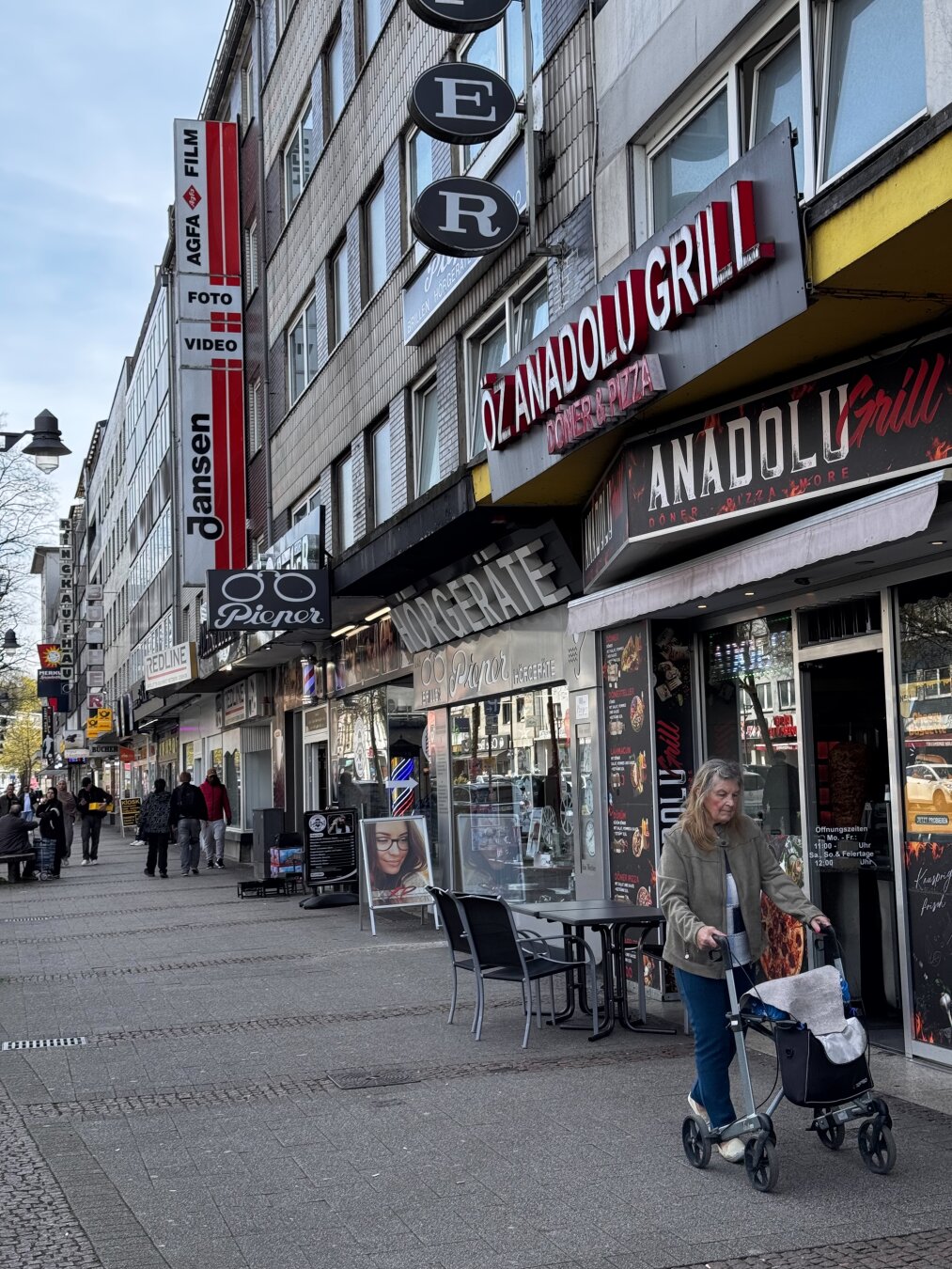 Eine belebte Straßenszene mit verschiedenen Geschäften, darunter ÖZ ANADOLU GRILL und Agga Foto. Eine Frau ist zu sehen, die mit einem Rollator unterwegs ist, während andere an Tischen im Freien sitzen. Schilder für Fotodienste und einen Friseur