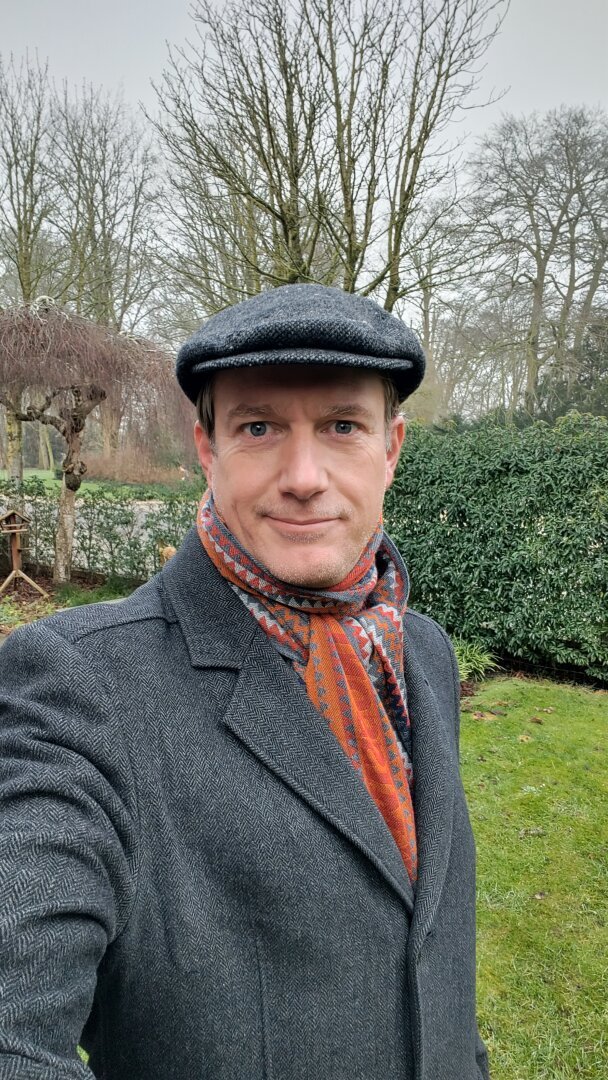 Man in overcoat, colourful shawl and cap.