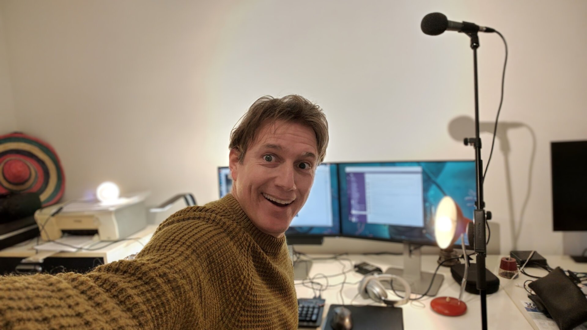 Man looking into camera in front of two computer screens, microphones and lights