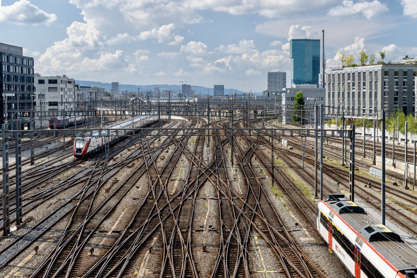 A tangle of metal train tracks cross all over the ground off into the horizon. Mid-rise buildings line the sides of the train tracks. It’s a sunny day with fluffy clouds.