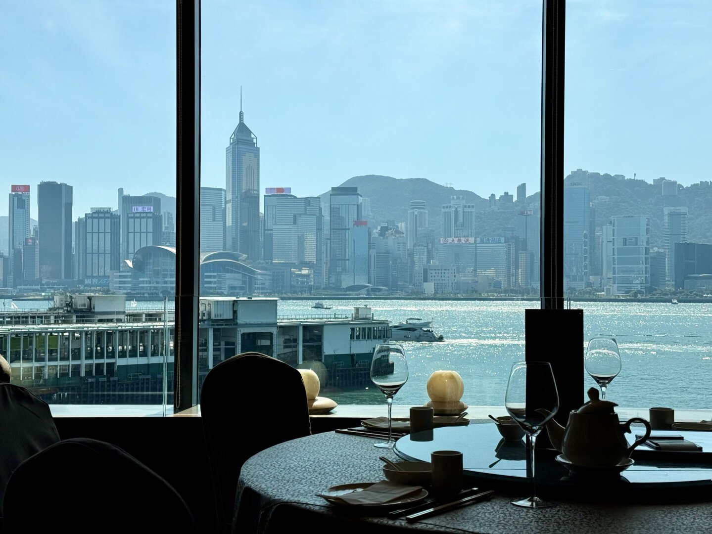 A photo of a dining room exposed for the bright daylight skyline and harbor outside. The dining room is dark cloth slipcovered chairs with white sateen tablecloths. Wine glasses, tea pots, a lazy Susan and several place settings with pairs of chopsticks and paper napkins.