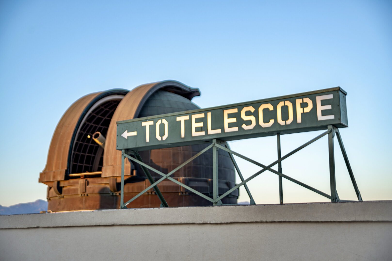 A photo of the large “TO TELESCOPE” metal sign at the roof level of the Griffith Observatory. The left side of the sign has an arrow pointing to the left. Right behind the sign on the left side of frame is the observatory telescope dome, open, showing the telescope lit by the setting sun behind camera. The sky is blue and featureless.