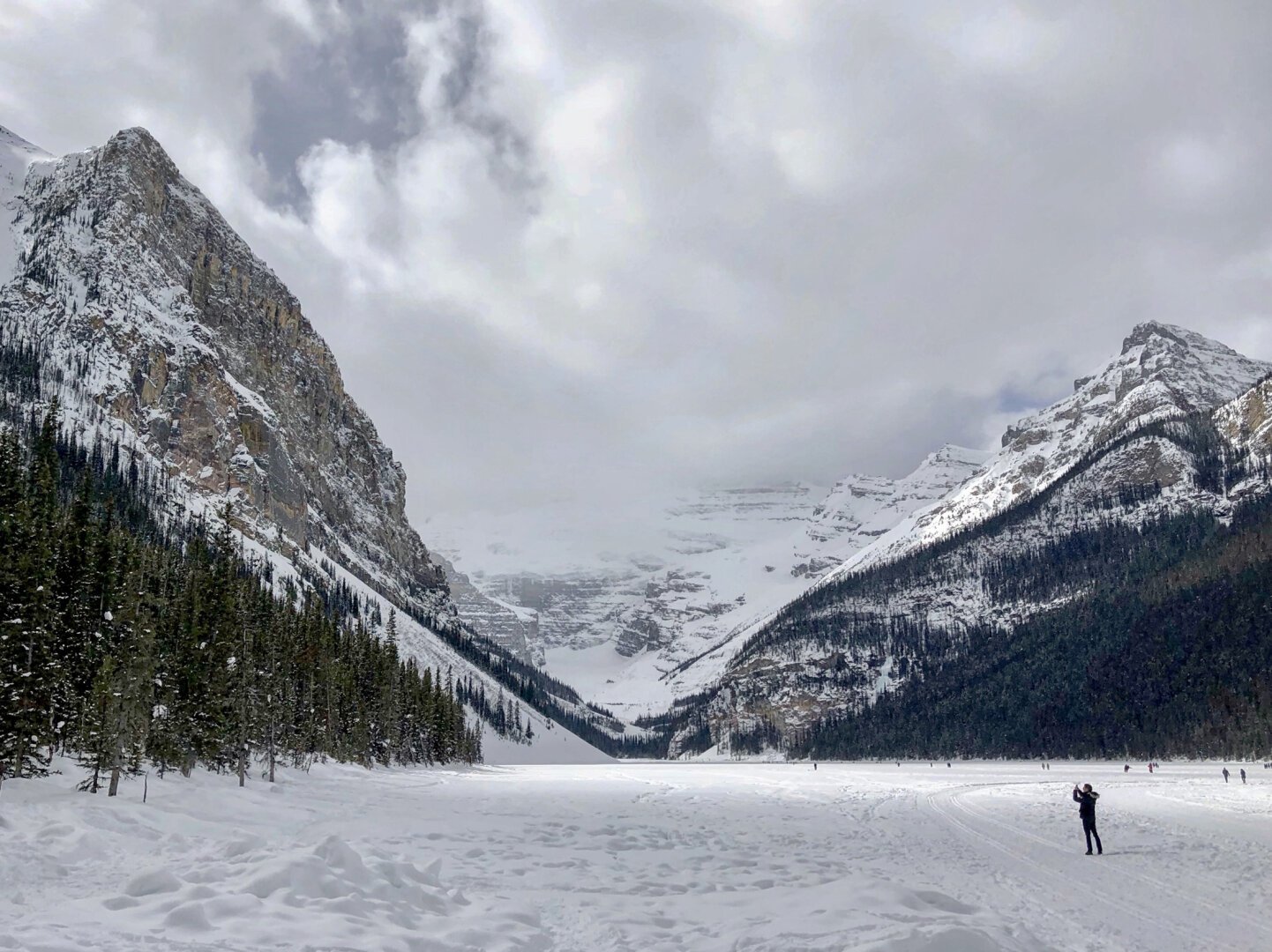 A photo of Lake Louise in the winter. The lake is frozen and covered in snow. The mountains form a V-shape in the center of frame. A figure in the lower right of frame is taking a photo.