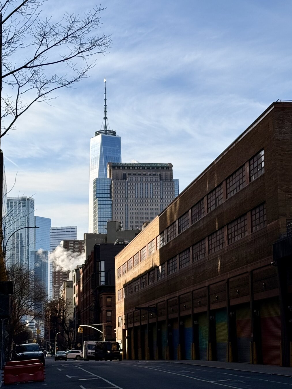 A photo of One World Trade Center from a few miles away tucked behind some other buildings and a warehouse with colorful doors that’s mostly in shadow. Wisps of steam in the sun. Parked cars line the street but no one is on the road.