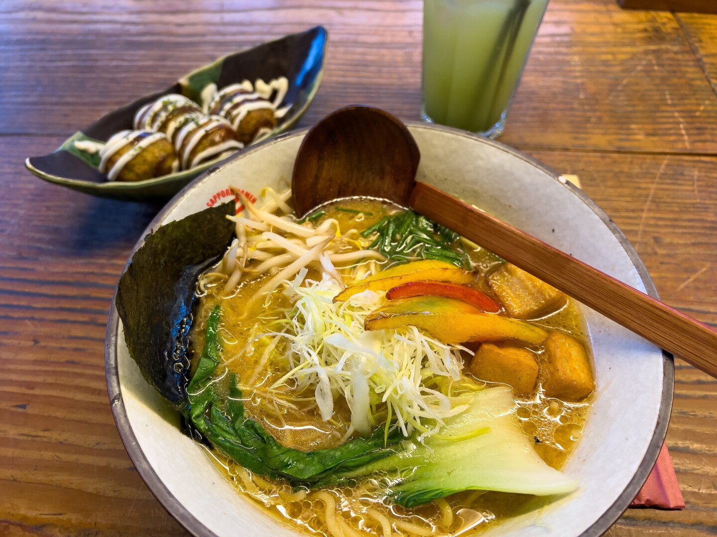 Japanische Ramen Nudelsuppe mit einem Löffel darin, daneben stehen Bällchen in einer Schale und kalter Matcha Grüntee in einem Glas.