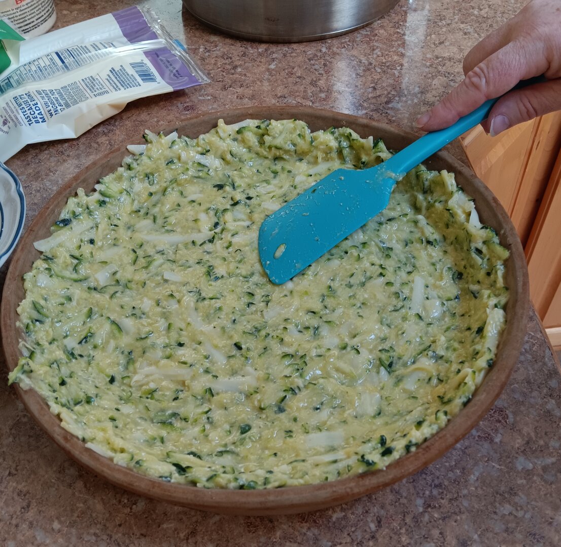 A hand uses a blue spatula to spread a yellow-and-green mixture across a round pan sitting on a brown counter. Various ingredients and kitchenware are visible in the background.