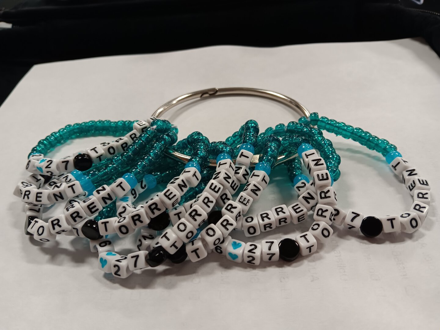 A pile of kandi bracelets strung through a silver ring sits on a white paper. Each reads "26 27 Torrent" with a round black 'puck' bead between 27 & Torrent, a white bead with a blue heart between the 26 & 27s, blue glow accent beads on either side of the text, and sparkly teal mini-pony beads filling out the rest of the bracelet.