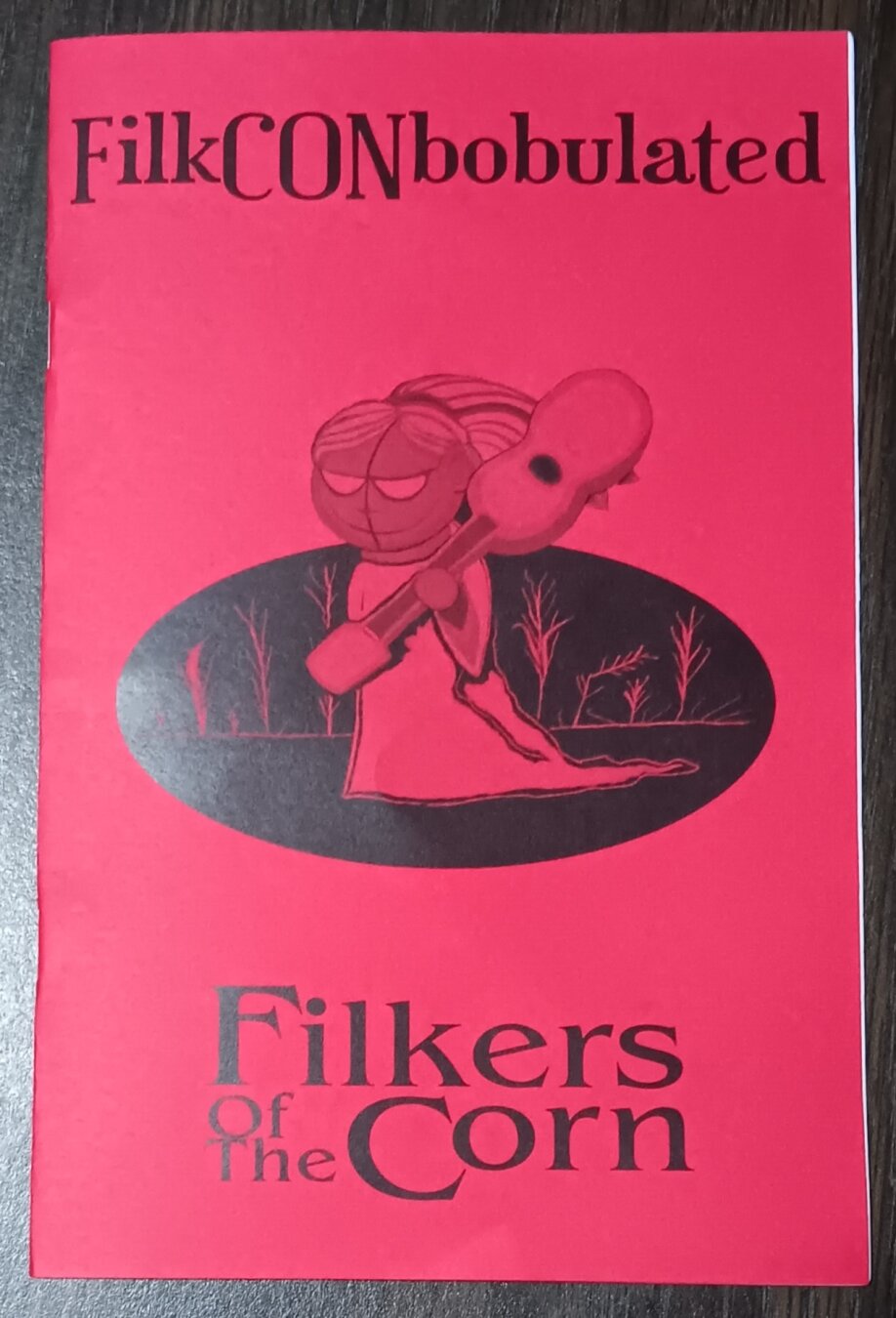 A red program book cover with black printing reads "FilkCONbobulated - Filkers of the Corn" around an image of a creepy doll in a floor-length gown walking through a sparse cornfield with a guitar-shaped object over her shoulder.