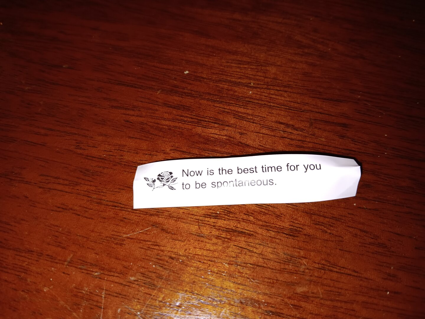 A white fortune slip on a brown wood tabletop reads "Now is the best time for you to be spontaneous."