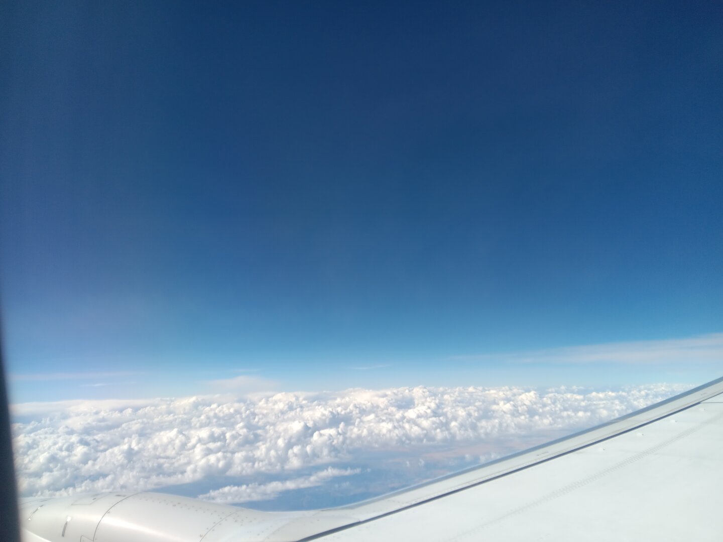 A bright blue sky above a highly-textured cloud layer, as seen from an airplane window. A bit of the wing can be seen at the bottom of the photo.