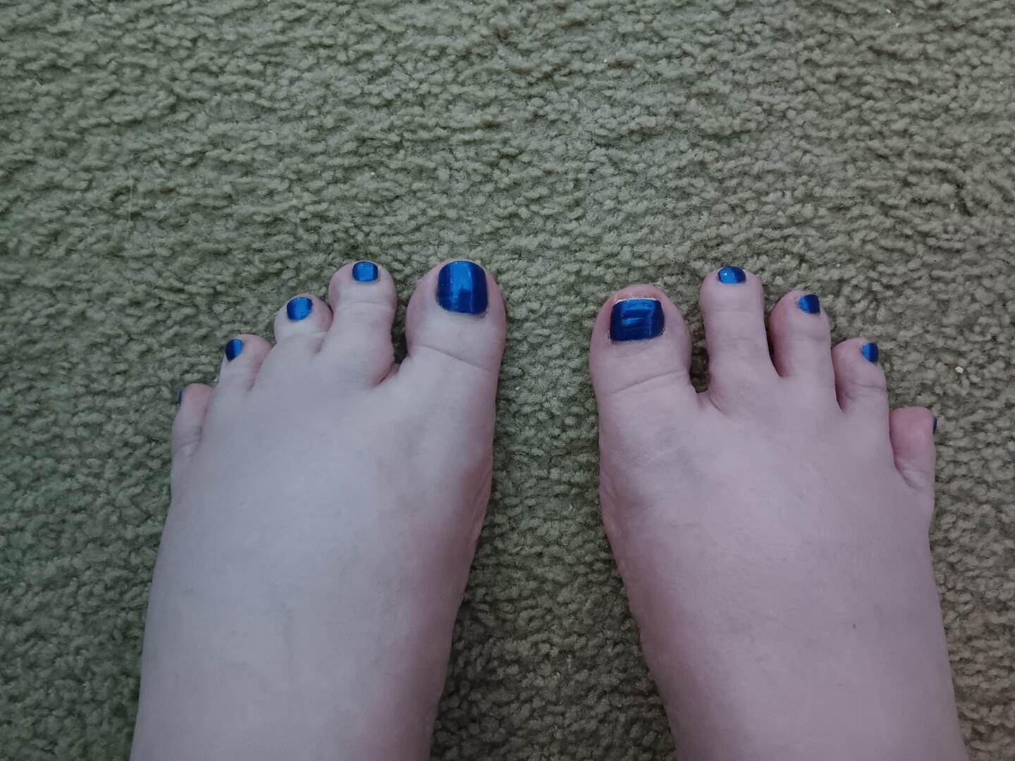 A pair of pale fair-skinned feet with bright cobalt-blue toenails sits on beige carpeting.