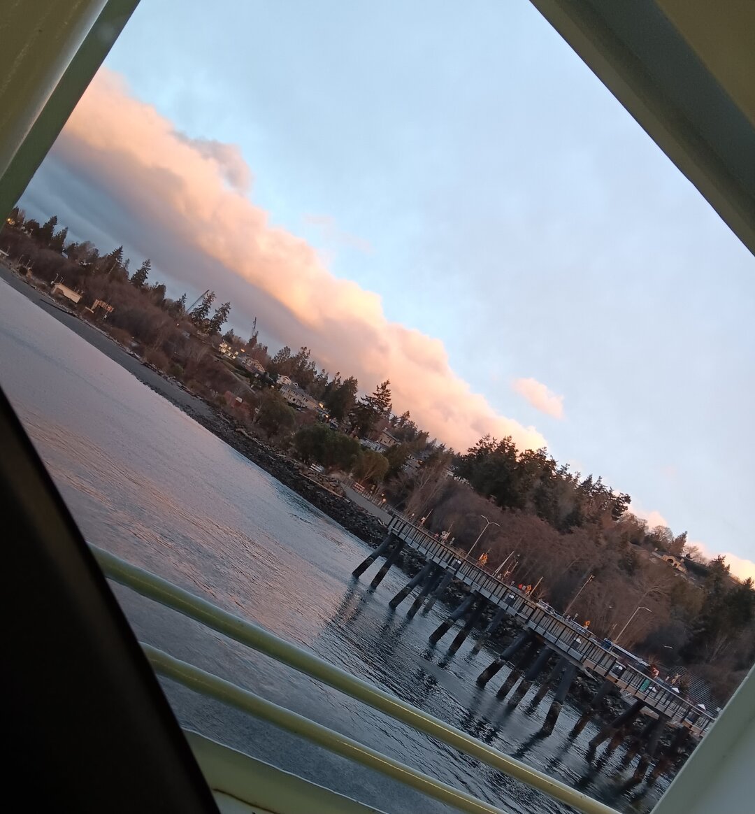 View from a ferrywindow showing a pier into the sound, with and and trees further back. Low clouds catch enough sunset to reflect lilac and peach tones, with light blue sky above.