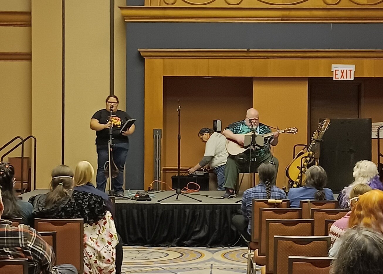 A stage in a hotel ballroom, with a short femme-appearing person singing into a mic on the left and a large man seated playing a guitar behind a mic on the right. A woman in a light shirt bends over a control board behind the stage, and bits of the audience are visible in the foreground.