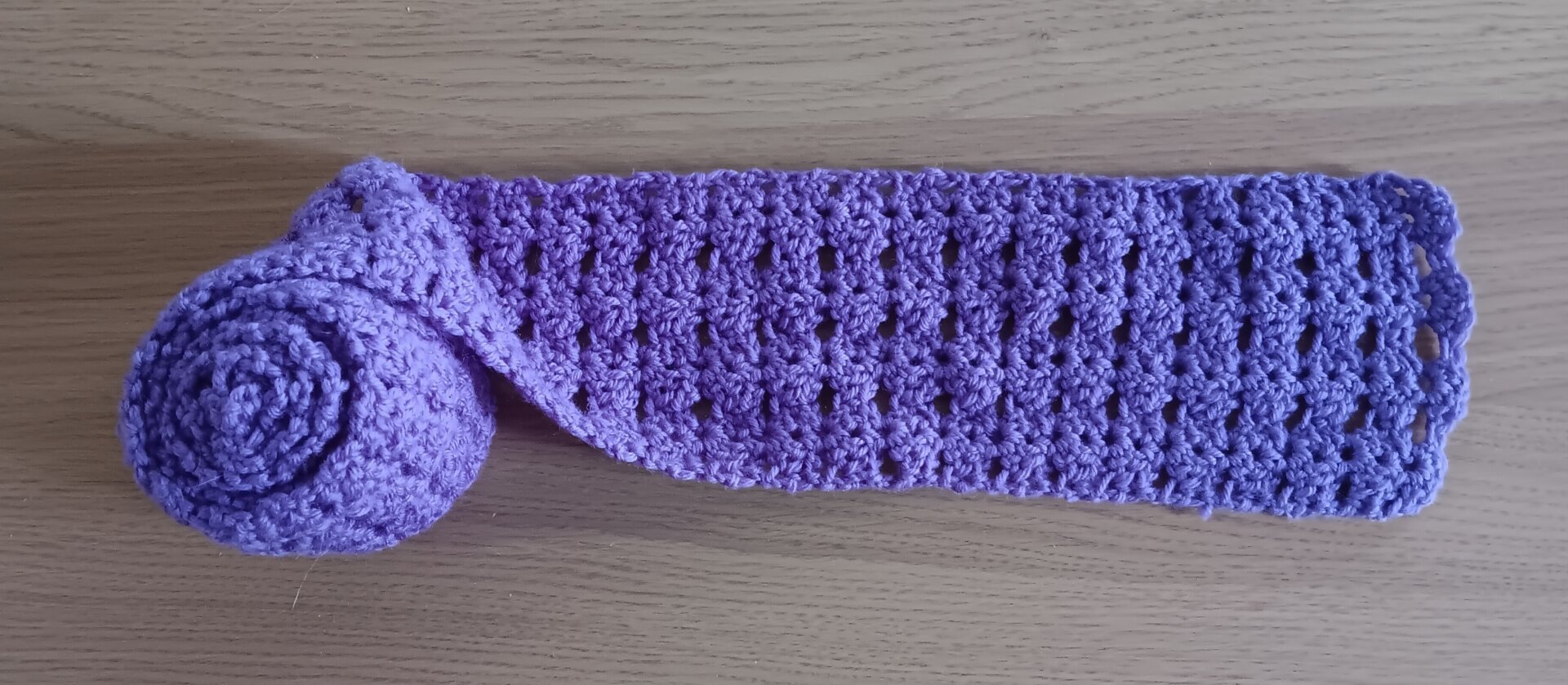 A foot or so of a crocheted purple scarf has been  stretched out from a rolled-up section on top of a pale woodtoned surface so the patterning shows.